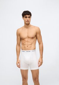Mužský model s krátkými tmavými vlasy, oblečený v bílých boxerkách Calvin Klein, stojící na jednoduchém světle šedém pozadí.