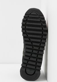 Semelle de chaussure noire avec de profondes rainures horizontales et le numéro de taille 37 visible au talon, montrée sur un fond blanc en angle.