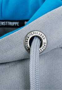 Gros plan sur le tissu gris d'un sweat à capuche avec un cordon bleu clair passant dans un œillet en métal gravé "SPITZBUB".