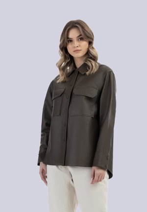 Jeune femme aux cheveux ondulés porte une veste en cuir marron foncé boutonnée avec des poches avant et un pantalon clair, debout devant un fond uni.