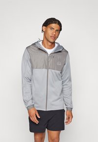 Sivi zip-jakna s kapuljačom, s lakšim sivim panelom preko prsiju. Glatka tekstura tkanine i logo detalj na lijevoj strani.
