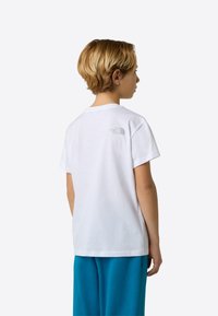 Biele bavlnené tričko s krátkymi rukávmi, na zadnej strane s malým sivým logom. Tričko je nosené s modrými teplákmi.