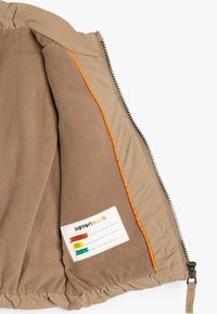 Interior de chaqueta acolchada Tan con costuras naranjas, forro de vellón suave y una etiqueta de cuidado con espacios en blanco para detalles de personalización.