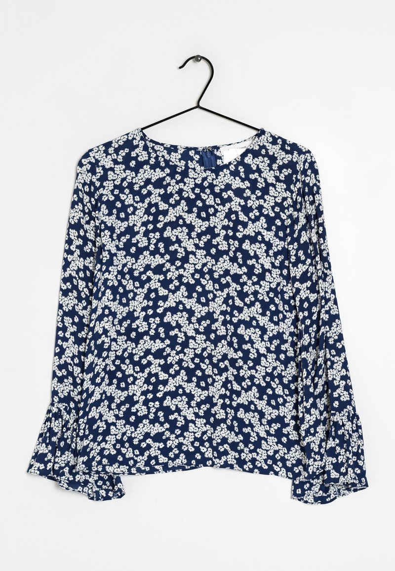 Blouse bleu marine à manches longues avec motif floral blanc, encolure ronde et poignets légèrement évasés, suspendue à un cintre en fil noir.