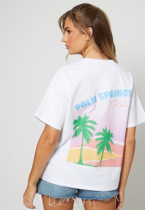 Femme portant un T-shirt blanc avec un imprimé coloré de Palm Springs en Californie et un short en denim, de profil contre un fond uni.