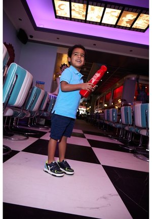 Kind in een lichtblauw polo shirt en donkere shorts, met een rode speelgoedfles in de hand, staat op een zwart-wit geblokte vloer in een diner.