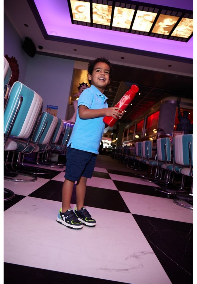 Un enfant portant un polo bleu clair et un short foncé, tenant une bouteille de jouet rouge, se tient sur un sol en damier noir et blanc dans un diner.