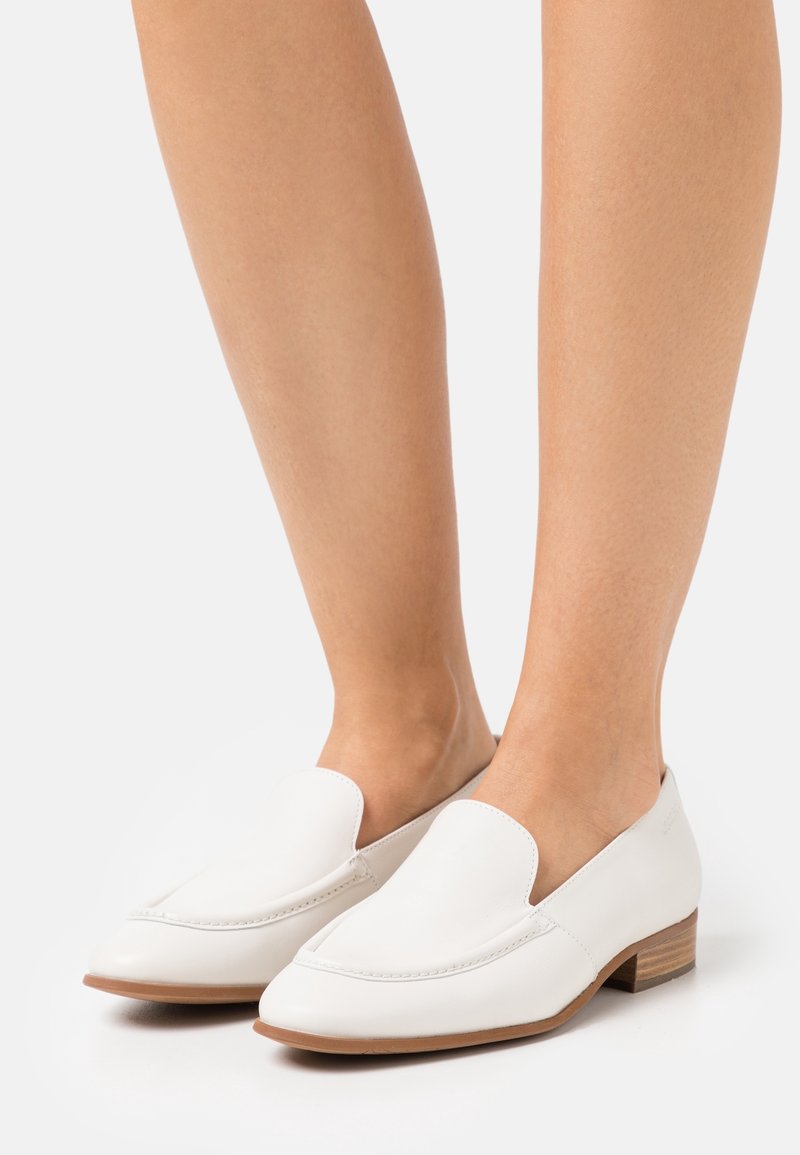 Pieds portant des mocassins blancs en cuir à enfiler avec de petits talons en bois sur un fond clair.
