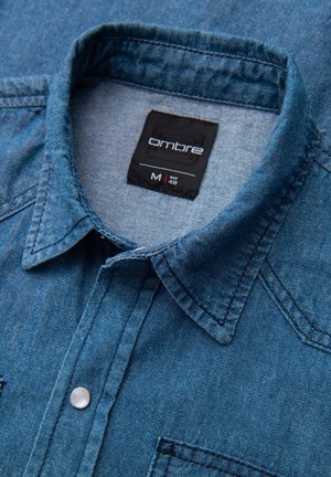 Camicia di jeans in tonalità blu scuro, con colletto appuntito, taschino sul petto e chiusure con bottoni a pressione argento. Etichetta con la scritta "Ombre M 48."