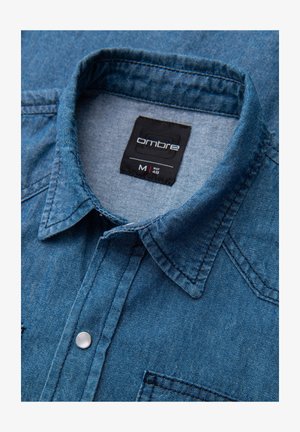 Jeanshemd in tiefblauem Farbton, mit spitzem Kragen, Brusttasche und silbernen Druckknöpfen. Das Etikett trägt die Aufschrift "Ombre M 48".