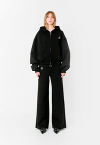 Sudadera negra de gran tamaño con cremallera frontal, que presenta un tirador circular y un acento de logo. Combinada con pantalones negros de pierna ancha y zapatos negros de punta.