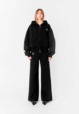 Sudadera negra de gran tamaño con cremallera frontal, que presenta un tirador circular y un acento de logo. Combinada con pantalones negros de pierna ancha y zapatos negros de punta.