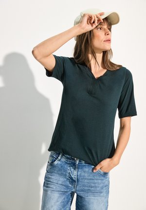 Dunkelgrünes T-Shirt mit kurzen Ärmeln und V-Ausschnitt aus strukturiertem Stoff, kombiniert mit hellblauen Denim-Jeans und einer weißen Kappe.