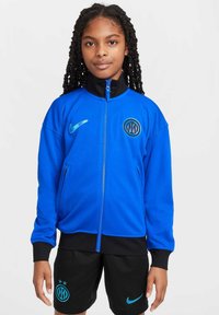 Giacca Nike blu con colletto e polsini neri, presenta un logo dell'Inter di Milano e tasche con zip. Indossata con pantaloni corti neri che mostrano il logo e dettagli.