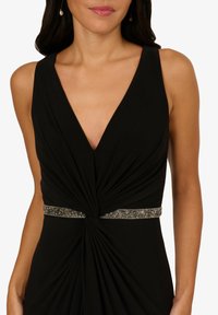 Černé večerní šaty s hlubokým výstřihem do V, nařaseným tělem a stříbrným korálkovým páskem v pase. Hladká textura a saténový vzhled.