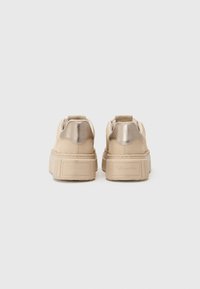Beige sneakers med metalliske detaljer, teksturert overdel, rund tå, tykk plattformsåle, og en trekkstroppe bak for enkel påkledning.