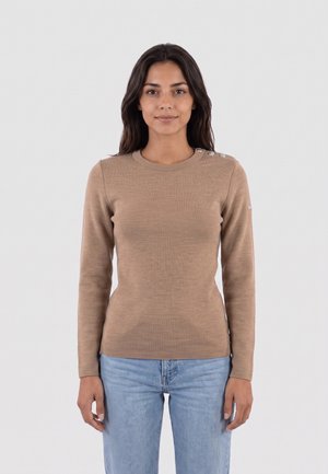 Donna con capelli scuri che indossa un maglione beige a maniche lunghe con bottoni sulla spalla e jeans azzurri, in piedi davanti a uno sfondo semplice.