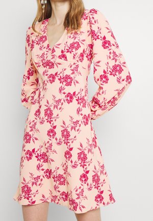 Woman wearing a light pink dress with red floral print and long puffed sleeves, standing against a plain light background.