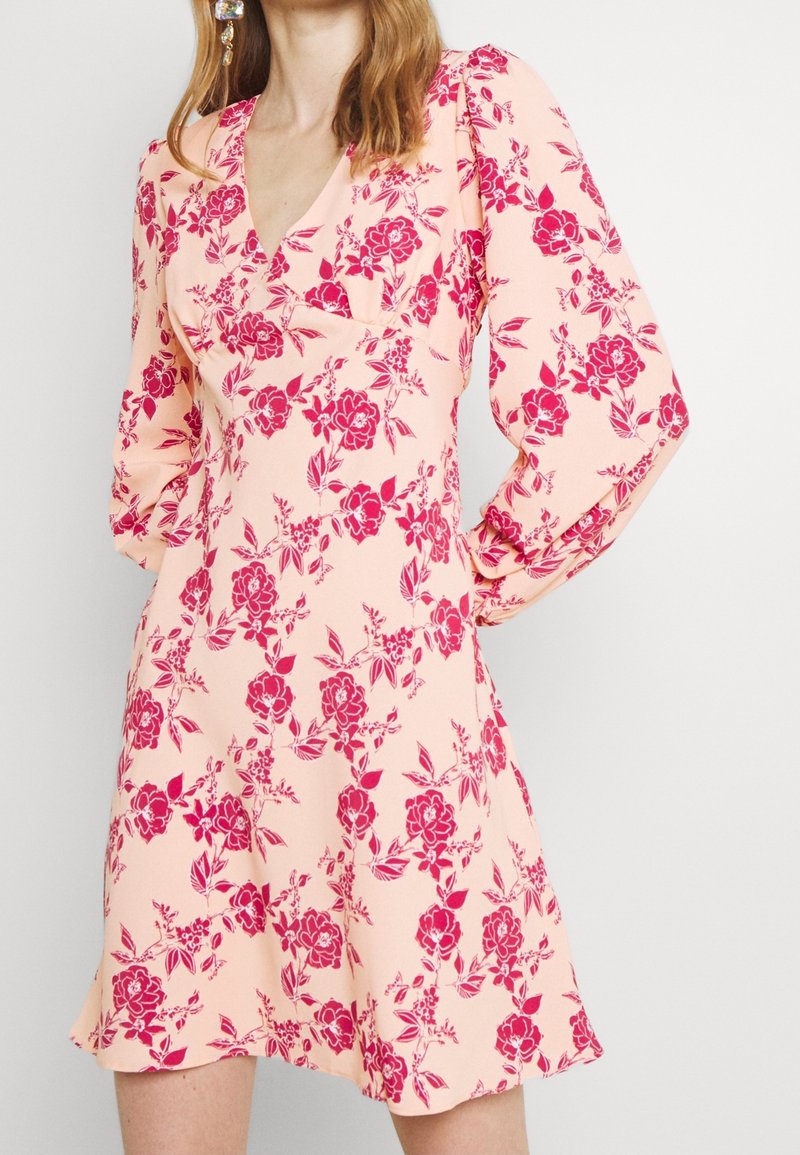 Femme portant une robe rose clair à imprimé floral rouge avec des manches longues bouffantes, debout devant un fond clair uni.