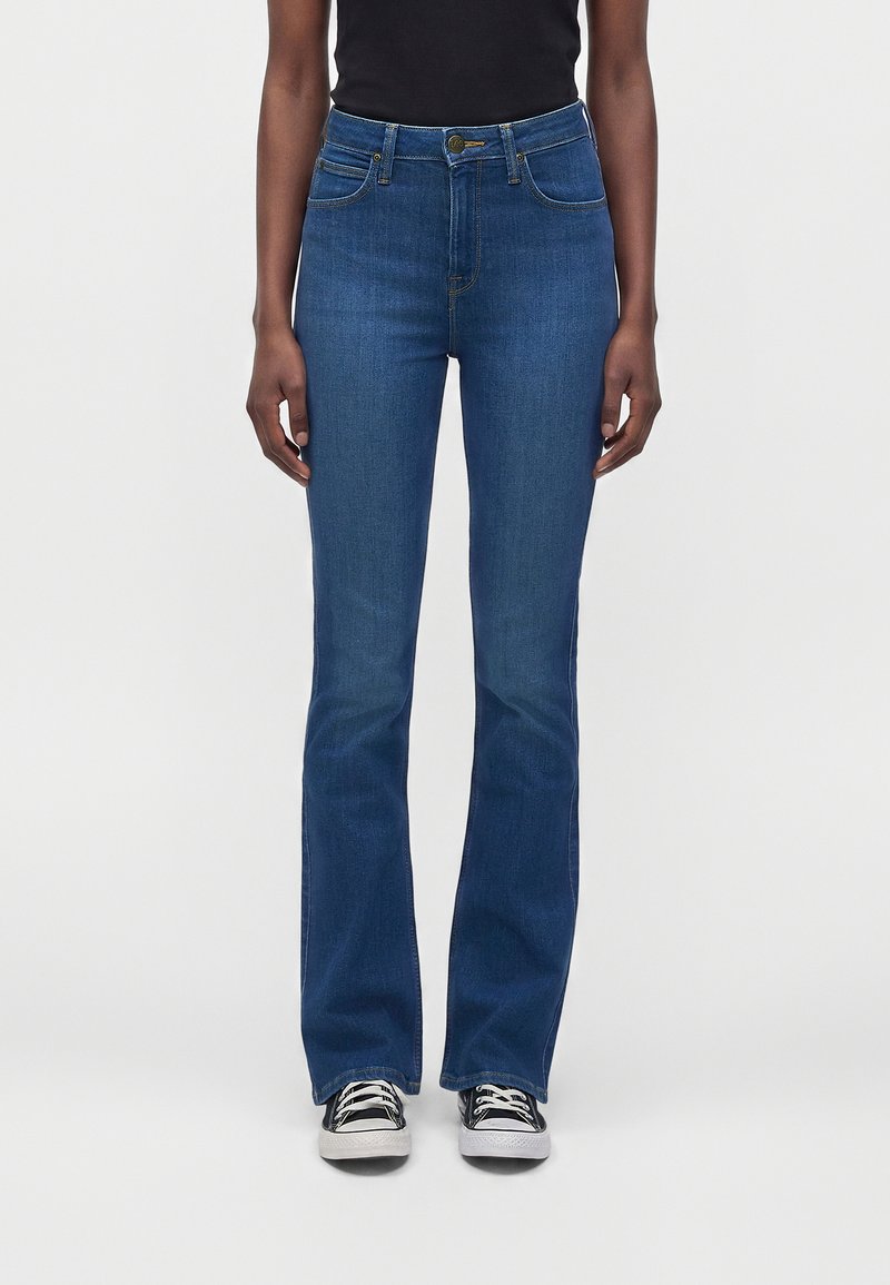 Femme portant un jean bootcut en denim bleu taille haute avec poches avant, débardeur noir et baskets noires et blanches, debout sur un fond blanc.