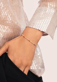 Mano que lleva una delicada pulsera de cuentas con pequeñas cuentas de colores, descansando sobre una tela negra, con una manga de lentejuelas en un tono beige claro visible.