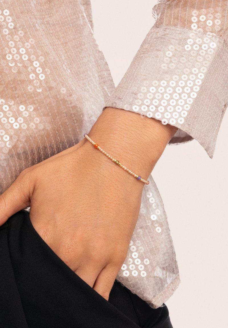 Mano que lleva una delicada pulsera de cuentas con pequeñas cuentas de colores, descansando sobre una tela negra, con una manga de lentejuelas en un tono beige claro visible.