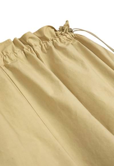 Tissu beige avec une ceinture élastique froncée et un cordon de serrage équipé d'une boucle ronde pour un ajustement réglable.