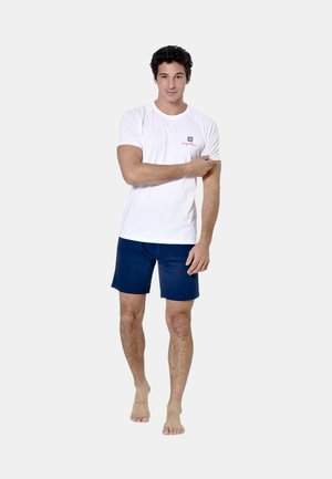 Homme debout pieds nus portant une chemise blanche à manches courtes et un short bleu marine, touchant son bras supérieur gauche sur un fond uni.