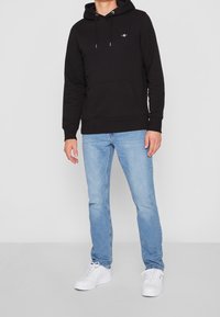 Sudadera con capucha negra y bolsillo frontal, combinada con jeans de color azul claro y zapatillas blancas, con un pequeño logo en el pecho.