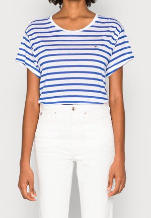 Personne portant une chemise à manches courtes rayée bleu et blanc rentrée dans un pantalon blanc taille haute, debout devant un fond uni.