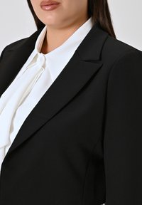 Zwarte op maat gemaakte blazer met scherpe revers, gemaakt van een soepele stof, gedragen over een witte blouse met een centrale knoop en zachte ruches.