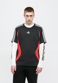 Μαύρο κοντομάνικο μπλουζάκι με κόκκινες πλαϊνές πάνελ, λευκό μακρυμάνικο πουκάμισο από κάτω και τρεις λευκές ρίγες στο ύψος των ώμων.