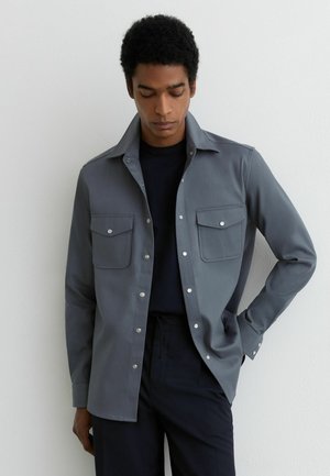 Mladý muž v sivej košeli na gombíky s náprsními vreckami, oblečený cez čierne tričko, stojaci s jednou rukou v vrecku oproti bielej stene.