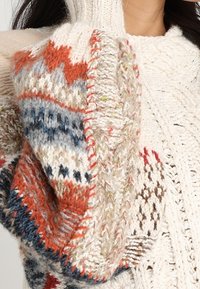 Hrubě pletený svetr s krémovou základní barvou a vícebarevnými geometrickými vzory v modré, červené a zelené na rukávech.