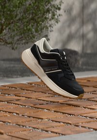 Μαύρο και εκρού αθλητικό παπούτσι με μαλακή σόλα, featuring υφή στην επάνω επιφάνεια, κλείσιμο με κορδόνια και λογότυπο "TOMMY JEANS" στο πλάι.