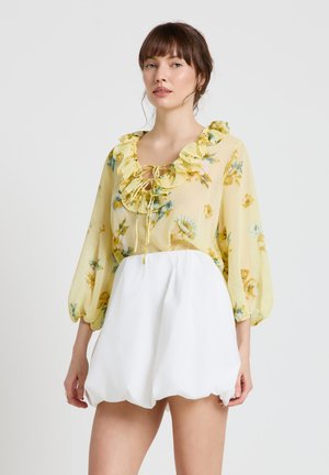 Jeune femme portant un chemisier jaune fleuri à volants avec manches trois-quarts et un short bouffant blanc taille haute sur un fond neutre.