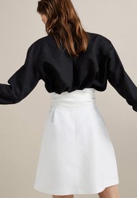 Donna con capelli castani che indossa un top nero a maniche lunghe e una gonna bianca a vita alta, girata di spalle con le braccia leggermente tese.