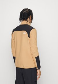 Jasno brązowa polarowa kurtka z czarnymi akcentami na ramionach. Posiada wysoki kołnierz oraz logo North Face z tyłu. Teksturowana tkanina.