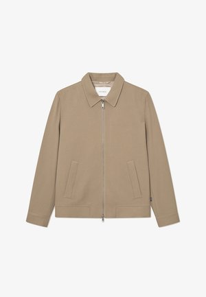 Veste beige avec zip sur le devant, col plat, deux poches avant et manches longues. Texture lisse et design minimaliste. Étiquette de la marque visible à l'intérieur.