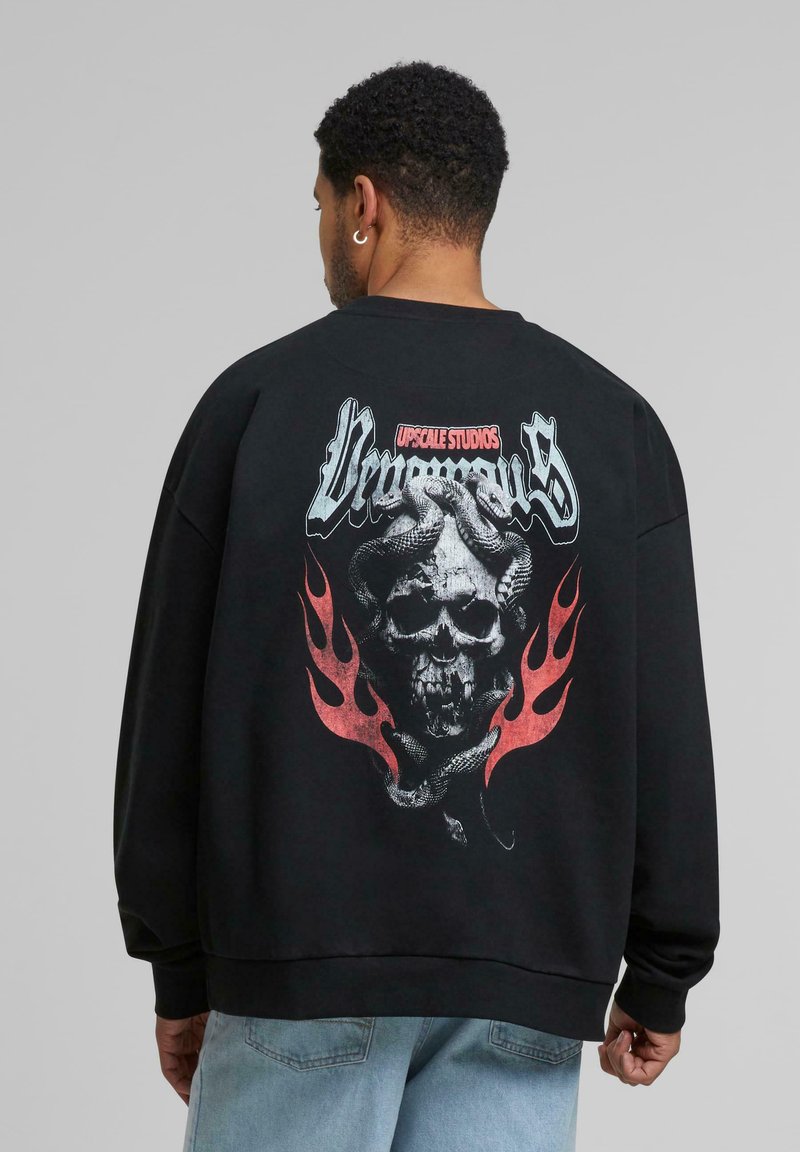 Czarny sweatshirt z długimi rękawami, z dużą grafiką czaszki oplecionej wężem, z czerwonymi płomieniami poniżej. Napis "Upscale Studios."