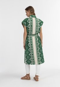 Abito verde e bianco a fantasia, con maniche corte, una cintura e un taglio al ginocchio, realizzato in un tessuto intrecciato che mostra motivi geometrici.