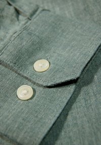 Detalle de una camisa de lino verde que presenta dos botones redondos color crema y una solapa en el cuello. La tela tiene una apariencia texturada.