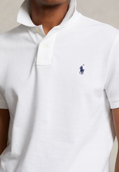Homme portant un polo blanc avec un col relevé et un petit logo joueur de polo bleu marine sur le côté gauche de la poitrine.