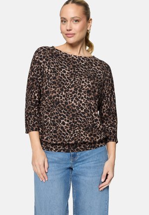 Frau mit hellem Teint und zurückgebundenen Haaren, die ein braun-schwarz gepunktetes Leopardenmuster-Oberteil und hellblaue Jeans trägt und vor einem einfarbigen Hintergrund steht.