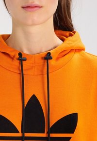 Närbild av en person som bär en orange hoodie med svarta snörningar och en stor svart Adidas-logotyp på framsidan.
