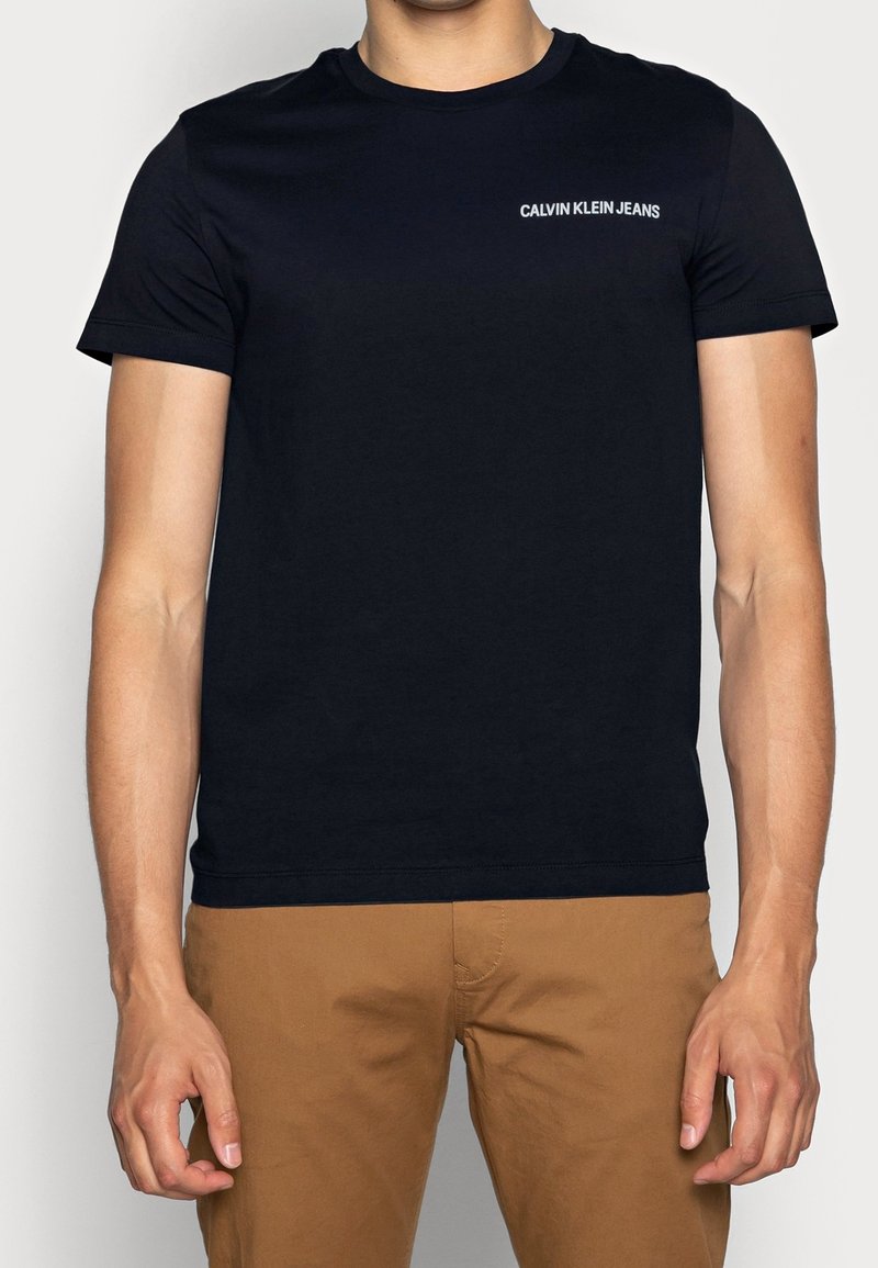 Musta puuvillapaita lyhyillä hihoilla, pyöreä kaula-aukko ja valkoinen "CALVIN KLEIN JEANS" -logo rinnassa, yhdistettynä vaaleanruskeisiin housuihin.