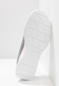 Grå atletisk sko med en texturerad ovandel, med en vit gummisula och en intrikat mönstring, placerad mot en vit bakgrund.