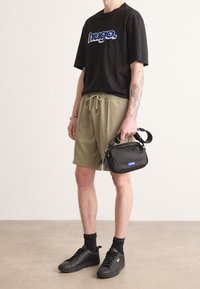 Musta t-paita, jossa sininen "hugo."-logo, oliivinvihreät shortsit, mustat lenkkarit ja pieni musta laukku sinisellä korostuksella ja olkahihnalla.