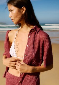 Bordeaux haakshirt gelaagd over een witte bikini, met grote knopen en een gouden kettinghalsketting, tegen een achtergrond van het strand.
