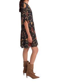 Kvinna i en svart knälång klänning med abstrakta repmönster och bruna mocka mitt-kalv klackstövlar, stående i profil.